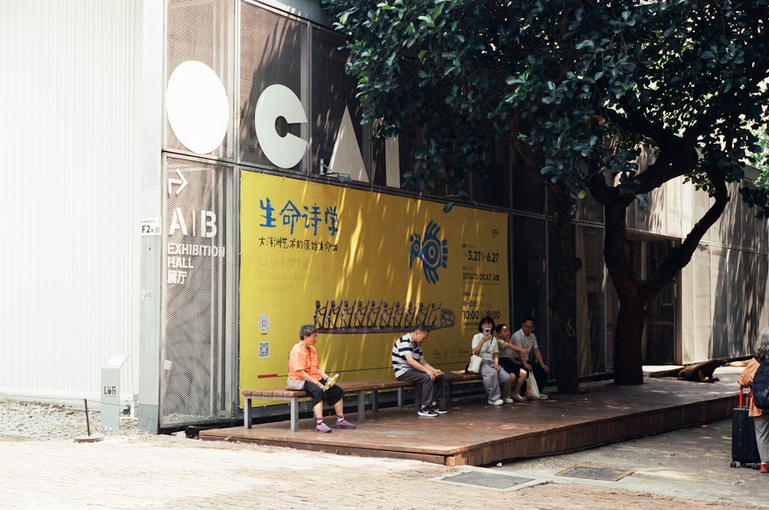 People sitting on benches under a tree near a building.
