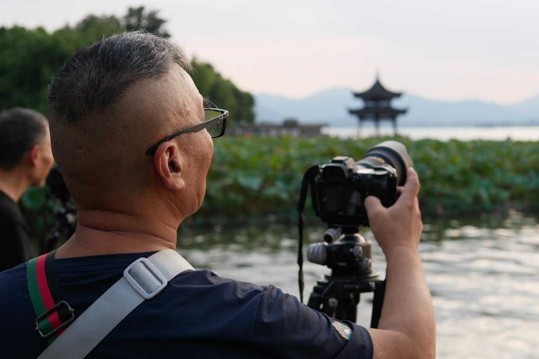 A man with glasses using a camera on a tripod by a lake