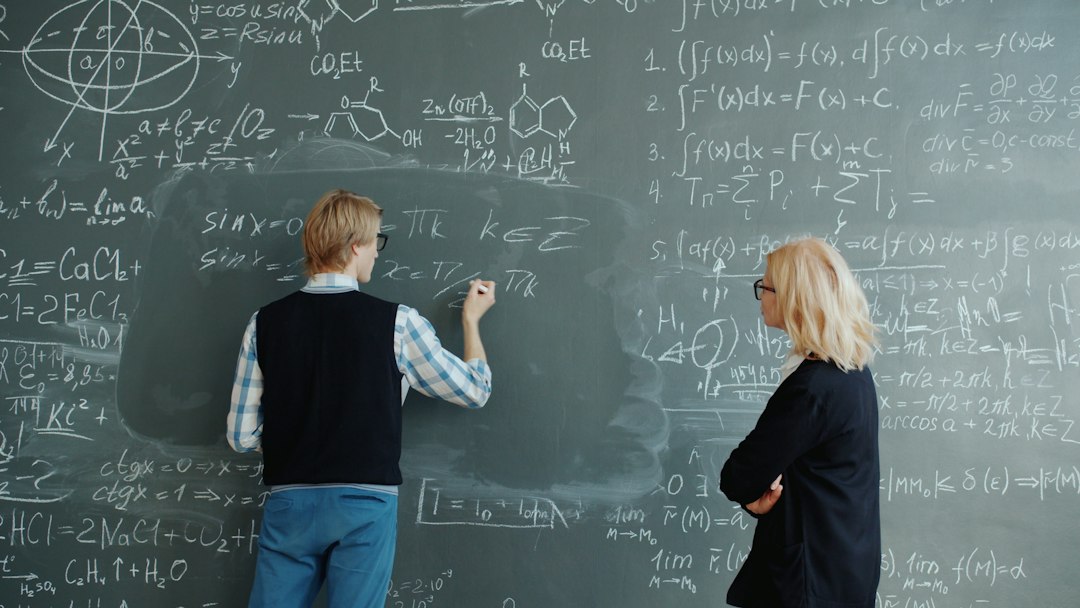 Student writes math equations on blackboard while teacher watches