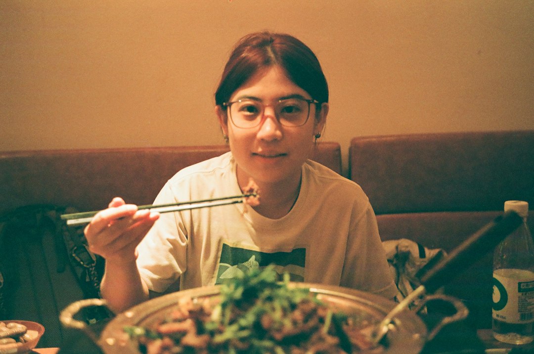 A person with glasses eating from a large shared dish with chopsticks in a restaurant